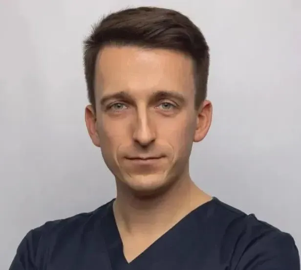 Mężczyzna z brązowymi włosami w granatowym fartuchu medycznym, stojący na białym tle.